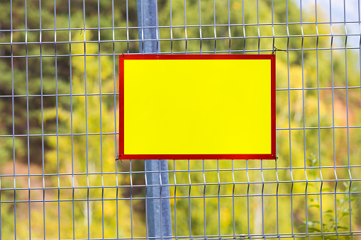 Empty yellow with red frame plastic sheet with mockup on metal wire fence. Blank plate for title. Place for text on the grid fence.Copy space for warning notification