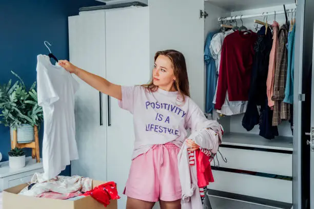 Photo of Woman selecting clothes from her wardrobe for donating to a Charity shop. Decluttering, Sorting clothes and Cleaning Up. Reuse, second-hand concept. Conscious consumer, sustainable lifestyle.