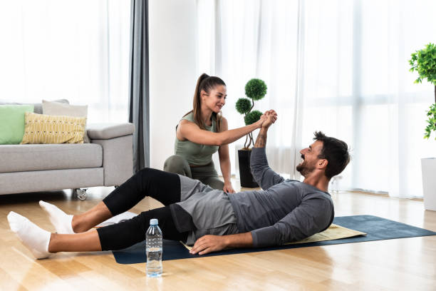 giovane istruttore di fitness e yoga che lavora a casa tua. donna terapista allenatrice educata sportiva che aiuta il giovane a recuperare nel suo appartamento e a casa in allenamento e allenamento per una vita sana. - yoga men male gymnastics foto e immagini stock