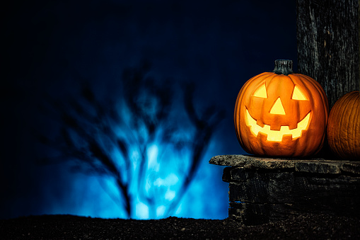 A glowing smiling pumpkin Jack O’ Lantern and pumpkin sitting on an old wooden porch with the glow of a rising full moon through a tree behind it.