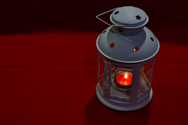 A close-up of a glowing candle inside a decorative lantern adorned with star patterns, creating a warm and magical Christmas ambiance. A blurred ceramic house in the background enhances the cozy holiday setting.