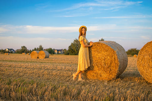 Young pretty woman in yellow dress and hat close up on the field background with haystacks at sunset time.