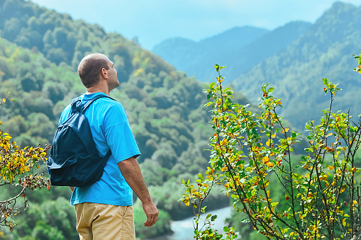 a male tourist with a backpack on his shoulders stands on the top of a mountain and admires a beautiful view of the nature of the mountains and the river below that opens before him