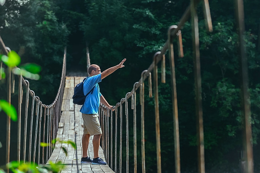 a male tourist with a backpack on his shoulders crosses a mountain river on a suspension bridge and waves to someone below