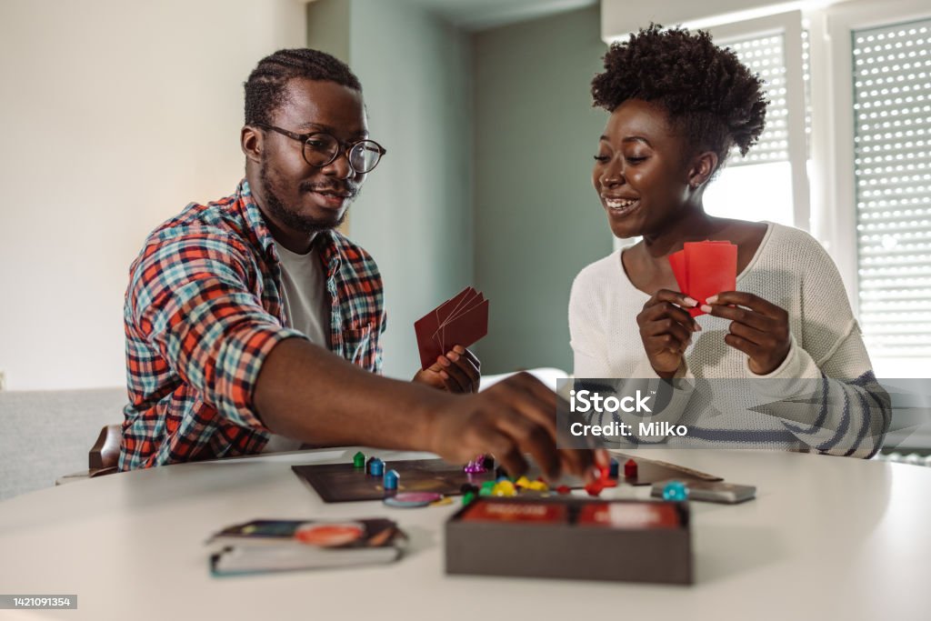 Young people enjoying at home and playing board game African-American young people sitting at the table in the living room and having fun while playing a board game Board Game Stock Photo