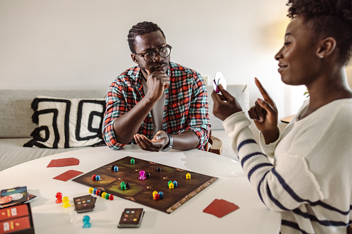 African-American young people sitting at the table in the living room and having fun while playing a board game