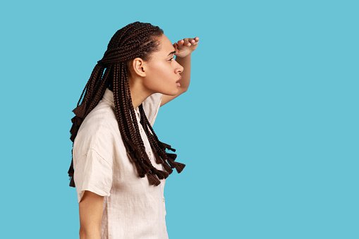 Side view of serious woman with black dreadlocks keeps hand near forehead, looks far away, searches something on horizon, wearing white shirt. Indoor studio shot isolated on blue background.