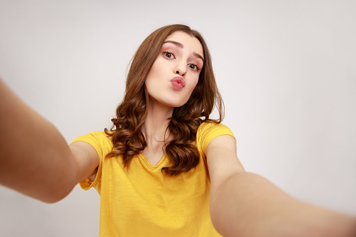 Portrait of lovely teenager girl in yellow casual style t-shirt taking selfie, looking at camera with pout lips POV, sending air kiss. Indoor studio shot isolated on gray background.