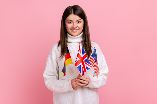 Beautiful young woman holds in hands British, German and American flags, polyglot studying languages, wearing white casual style sweater. Indoor studio shot isolated on pink background.