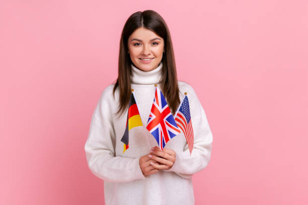 Beautiful young woman holds in hands British, German and American flags, polyglot studying languages Beautiful young woman holds in hands British, German and American flags, polyglot studying languages, wearing white casual style sweater. Indoor studio shot isolated on pink background. Teaching English Abroad stock pictures, royalty-free photos & images