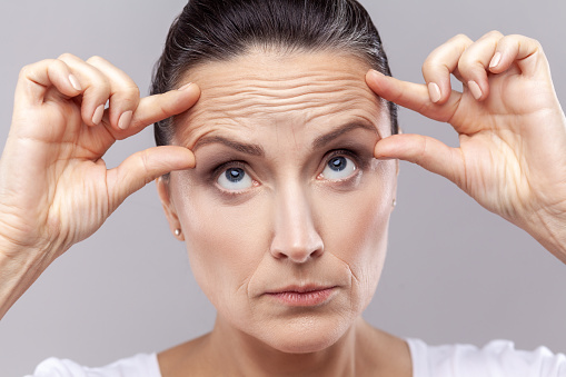 Closeup portrait of middle aged woman checking wrinkles, doing anti aging face yoga exercises to firm and tighten skin and relax muscles on her forehead. Indoor studio shot isolated on gray background