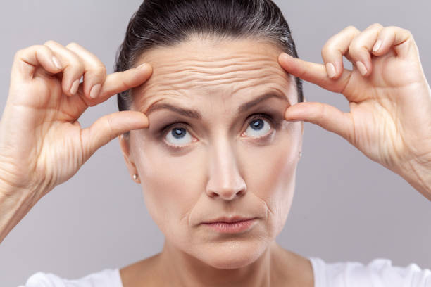 Woman checking wrinkles, doing antiaging face yoga exercises to firm and tighten skin. Closeup portrait of middle aged woman checking wrinkles, doing anti aging face yoga exercises to firm and tighten skin and relax muscles on her forehead. Indoor studio shot isolated on gray background wrinkled forehead stock pictures, royalty-free photos & images