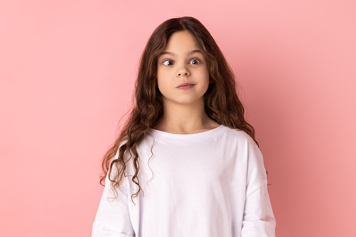 Portrait of little girl wearing white T-shirt crossing her eyes looking crazy and stupid, fooling around, having fun, vision problems. Indoor studio shot isolated on pink background.