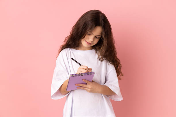 adorable little girl smiling and writing down interesting idea in notebook, making plans, to-do list - child thinking writing little girls imagens e fotografias de stock