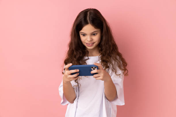Portrait of surprised little girl wearing white T-shirt using mobile phone with shocked positive expression, playing video game on cellphone. Indoor studio shot isolated on pink background.