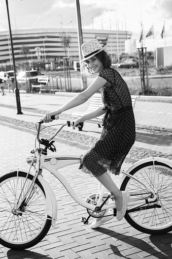 young curly hair woman in hat and dress cycling on bike at european city, sunset time, looking at camera, smiling, monochrome