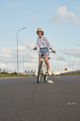 young woman cyclist in white blouse and hat moves on bicycle on road smiling