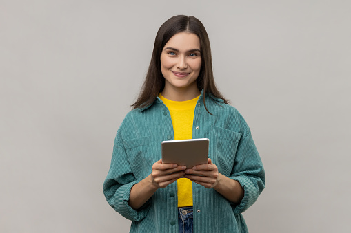 Woman with dark hair working online, using tablet for checking social networks, looking at camera with positive expression, wearing casual style jacket. Indoor studio shot isolated on gray background.