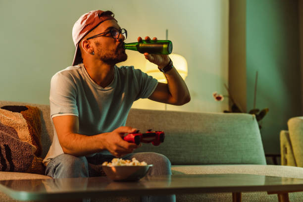 young man sitting on the sofa drinking beer and playing video games - after work beautiful people beer beer bottle imagens e fotografias de stock