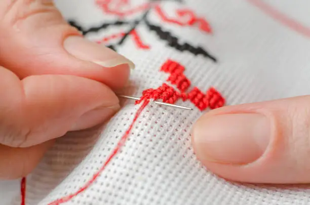Close-up of female hands cross stitching on a white canvas. A woman embroiders the national symbols of Ukraine with a cross.