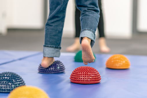 Several colorful squishy, spiky balls line the floor of a PT clinic as a child's feet walk across them.