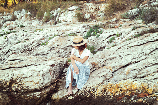 Girl in a straw hat sits on a rock by the sea on a sunny summer day.