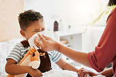 Flu, sick or cold child with parent sneezing, blowing and wiping runny nose while ill with covid virus, sinus and allergy symptoms in bed at home. Mother caring for stuffy and congested little son