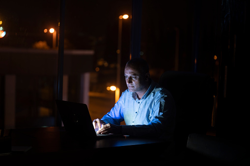 Photo of adult man using laptop computer in office during night. Shot in dark with a full frame mirrorless camera.