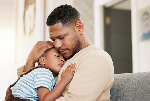 Sick, sleeping and tired child with caring father holding his son with a cold, flu fever or covid. Worried dad hugging his young boy on a living room family couch indoors feeling stress and worry