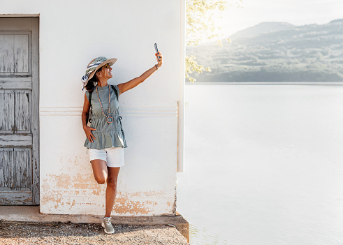 a woman with hat and scarf, is leaning on the facade of a house next to a lake and takes a selfie, while smiling. fun and nature concept.