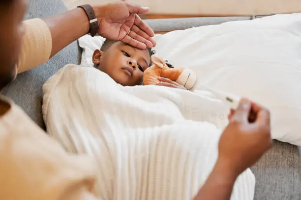 Cold, sick and covid child in bed, sad and ill with allergy and flu at home with his worried mother. Parents hand on son's forehead for his temperature as she checks his fever with a thermometer