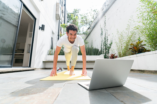 Happy Latin American man exercising at his home terrace and following an online fitness class on his laptop