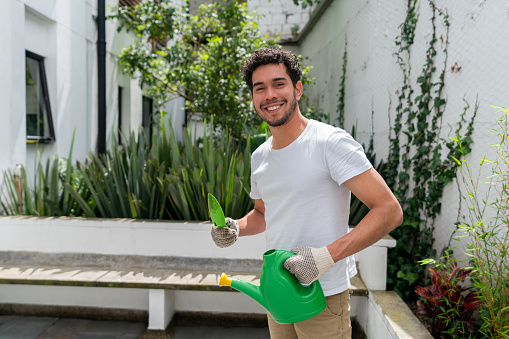 Happy Latin American man taking care of his home garden and watering the plants while looking at the camera smiling