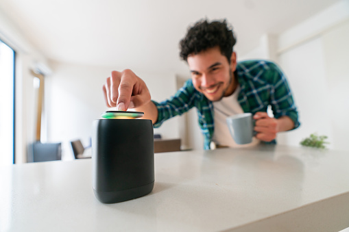 Happy Latin American man using a smart speaker at home while drinking a cup of coffee - virtual assistant concepts