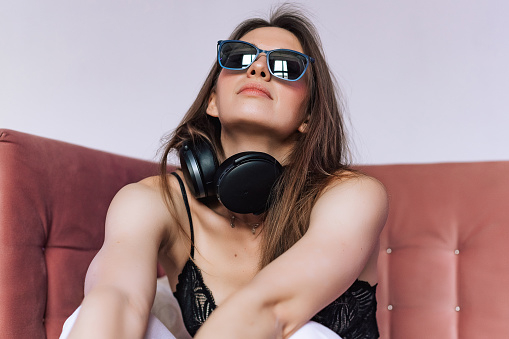 Female portrait of young woman indoors the room. Lifestyle photo in studio. Girl with eyeglasses on the sofa. Person using wireless headphones for music