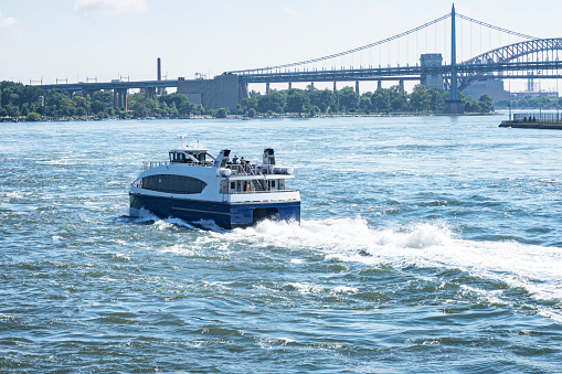 A New York City catamaran water ferry tour boat is moving tourist and commuter passengers across the rough, choppy East River water between the boroughs of Queens and Manhattan.