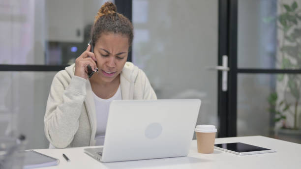 istock Angry African Woman Talking on Smartphone while using Laptop in Office 1421143060