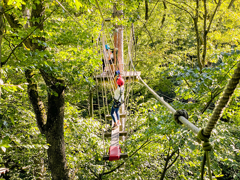 Rope park in wood forest.Happy school girl enjoying activity in a climbing adventure park on a summer day.Summer fun and sports for adventurous people.Teenager Girl walks a rope bridge between trees in an amusement park in safety gear and a helmet.