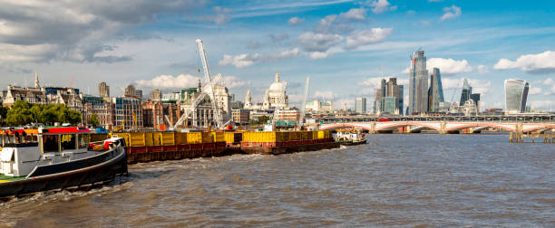 London city skyline,boats and barges drifting along the Thames river,on a warm summer afternoon,London,England,UK. Beautiful panoramic summer scene, depicting daily life on the iconic River Thames,at full tide,with blue skies and drifting clouds overhead. panoramic riverbank architecture construction site stock pictures, royalty-free photos & images