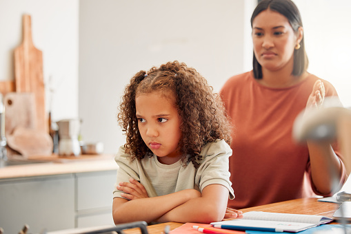 Unhappy, moody and angry little girl standing with arms crossed and looking upset while ignoring her mom. Upset daughter being scolded and reprimanded by her angry and disappointed mother at home
