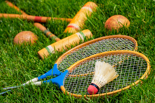 A low angle view of a real feathered shuttlecock sitting atop a pair of vintage wooden badminton rackets along with a pair croquet mallets and balls lying in the grass