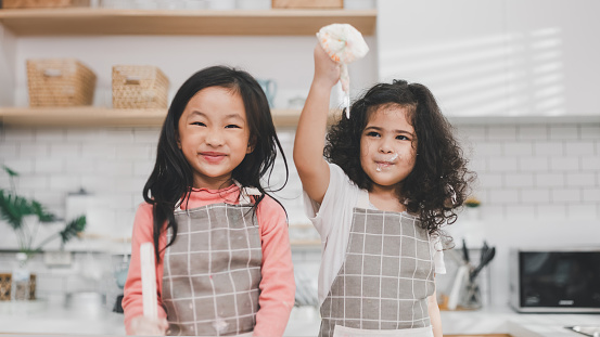 Children enjoy cooking in the kitchen. Happy Asian kids decorating the cake, bake cookies in the kitchen. Two cute little diversity girls play and learn trough the experience. Education concept.