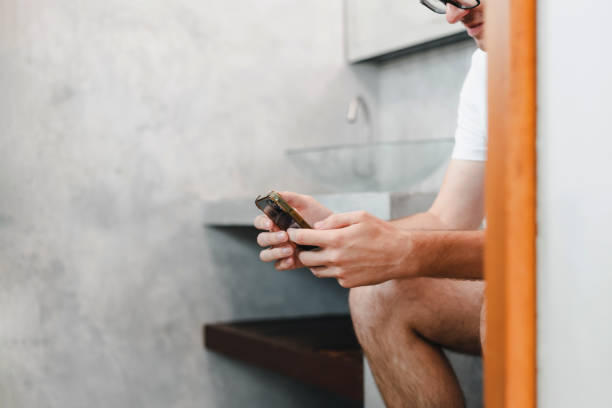 若い白人男性ゲーマーは、携帯電話のスマートフォンガジェットでオンラインでゲームをプレイトイレの便器に座っています。依存症依存症の習慣は、ソーシャルネットワークのメディア技� - mobile phone smart phone toilet water ストックフォトと画像