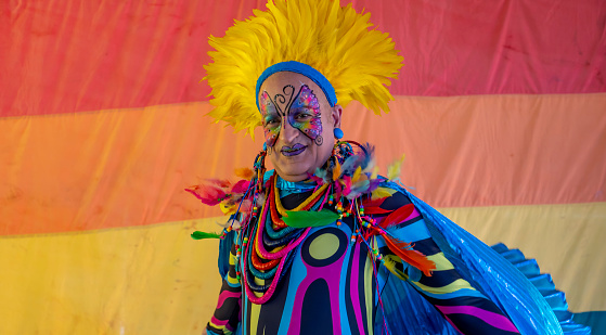 Lismore, NSW: August 28, 2022.\nMan in brightly coloured outfit dances in front of Gay Rainbow flag.\nLGBTQIA + is an initialism that stands for lesbian, gay, bisexual, and transgender. In use since the 1990s, the initialism, as well as some of its common variants, functions as an umbrella term for sexuality and gender identity. This community in Lismore NSW calls itself Tropical Fruits and has a number of celebrations throughout the year. This event happened after three years of cancellation due to covid and the worse floods in history. It is a free, largely outdoor event which involves dressing up and having a dog show.