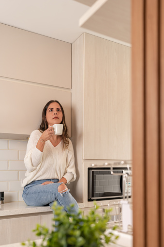 Vertical photo of a woman sitting at the counter in a new and modern kitchen drinking coffee