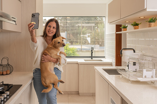 Woman taking a selfie with a mobile while holding a dog in the kitchen of a new house