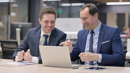 Middle Aged Businessman and Young Colleague Celebrating While using Laptop