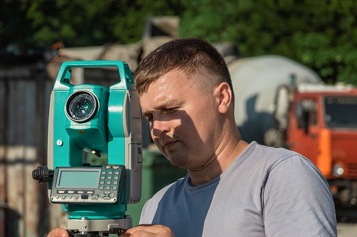 A male construction worker at an outdoor construction site. A surveyor with a total station at work on a sunny day. The topographer is located next to the total station.
