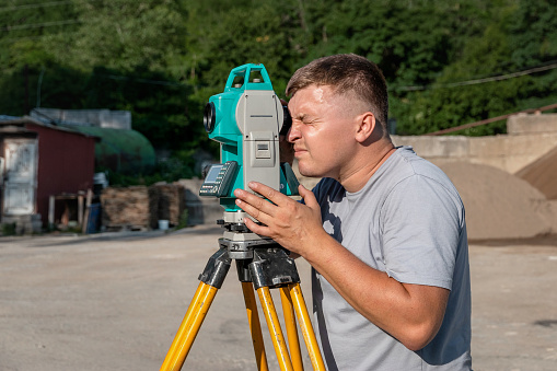 Portrait of a young man next to a total station in the open air. A surveyor with a total station at work on a sunny day. A male construction worker on an open construction site.