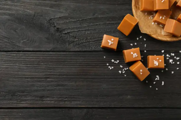 Yummy caramel candies and sea salt on black wooden table, flat lay. Space for text